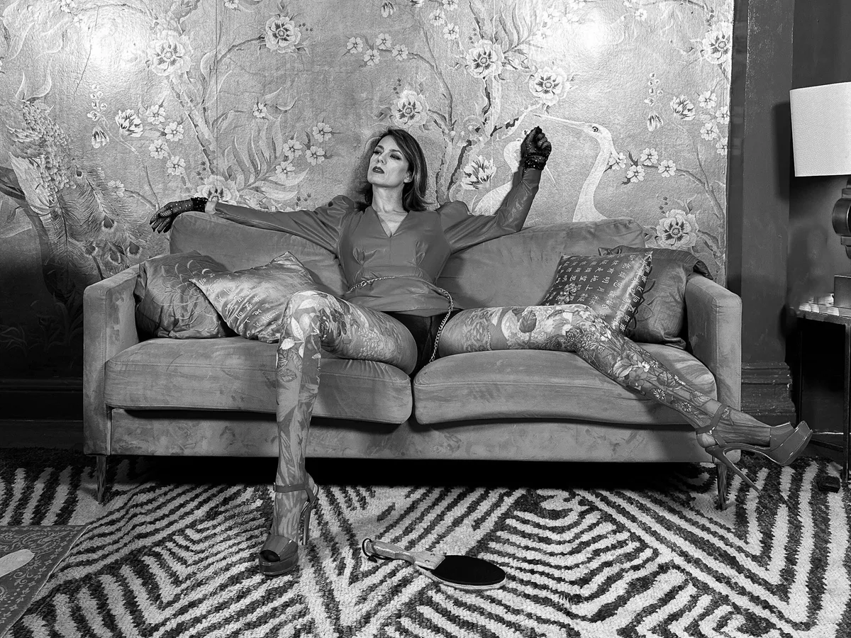 NYC Dominatrix Viktoria Sway in black and white, sprawled across a velvet sofa with both arms along its back, in sheer gloves, patterned tights and platform heels, a leather paddle lying untouched on the rug in front of her