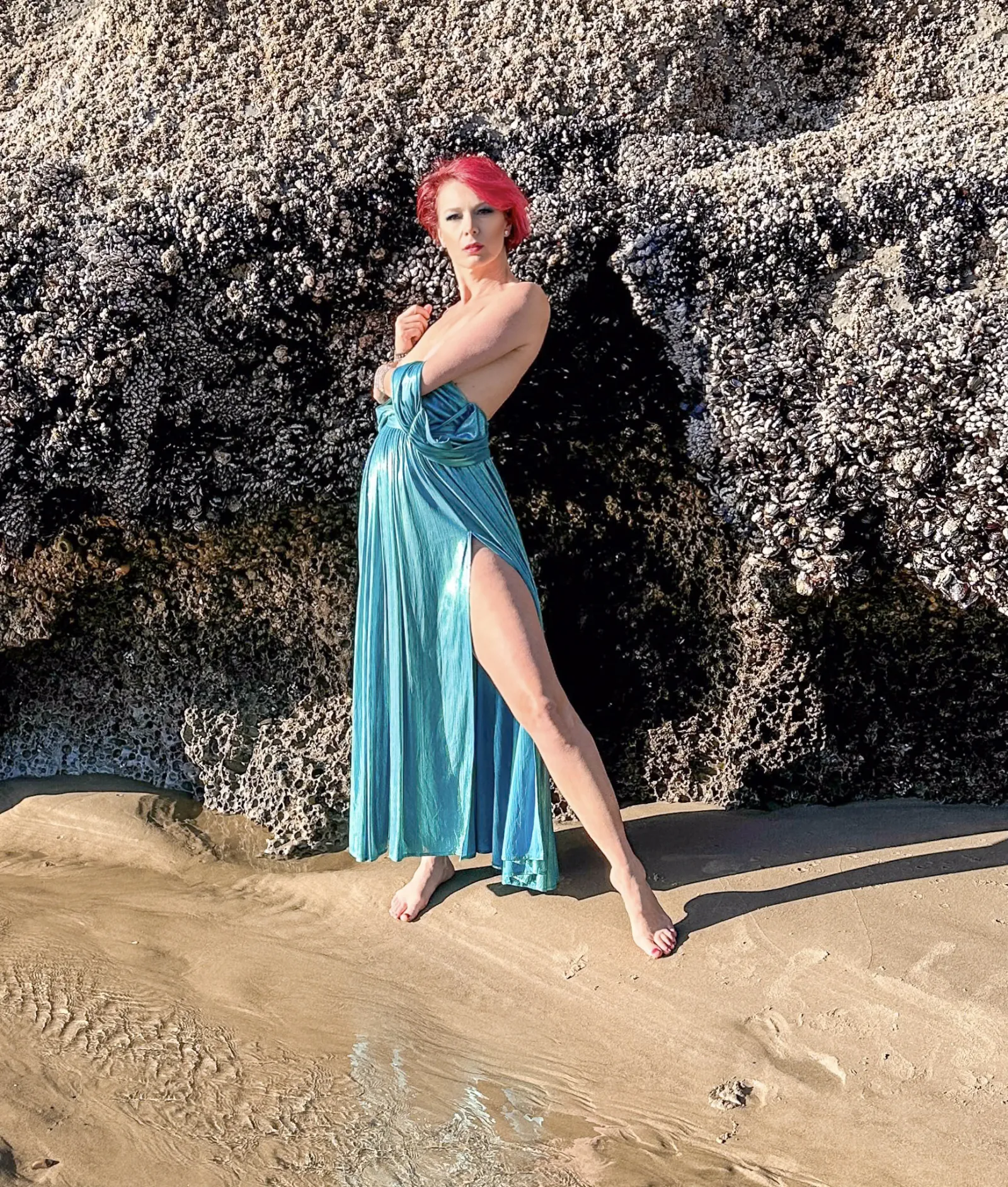 Viktoria poses in a flowing teal gown against barnacle-covered rocks on a sunlit beach, red hair catching the light