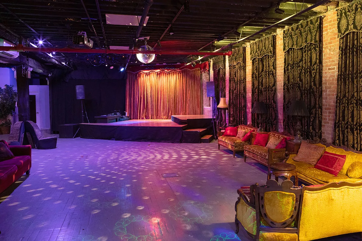 The Gemini and Scorpio Loft in Brooklyn: a long room with a low stage and curtain at one end, velvet sofas and lamps along an exposed brick wall, and an open wooden dance floor