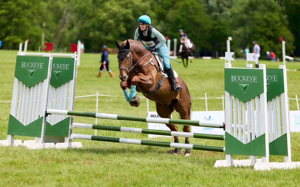 Viktoria smiling and waving while seated on a bay horse in equestrian gear outside a stable.