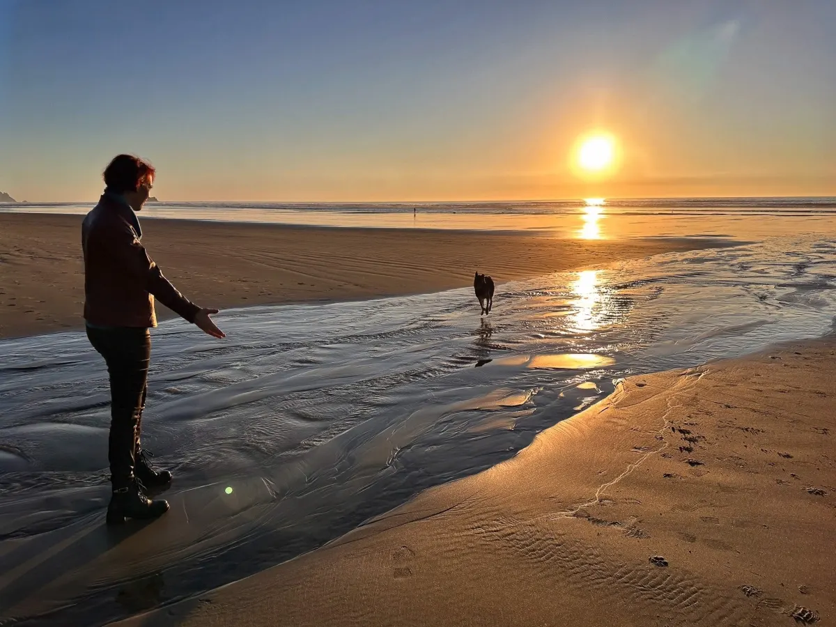 Viktoria reaching toward a dog at sunset on the beach, atmospheric full-bleed