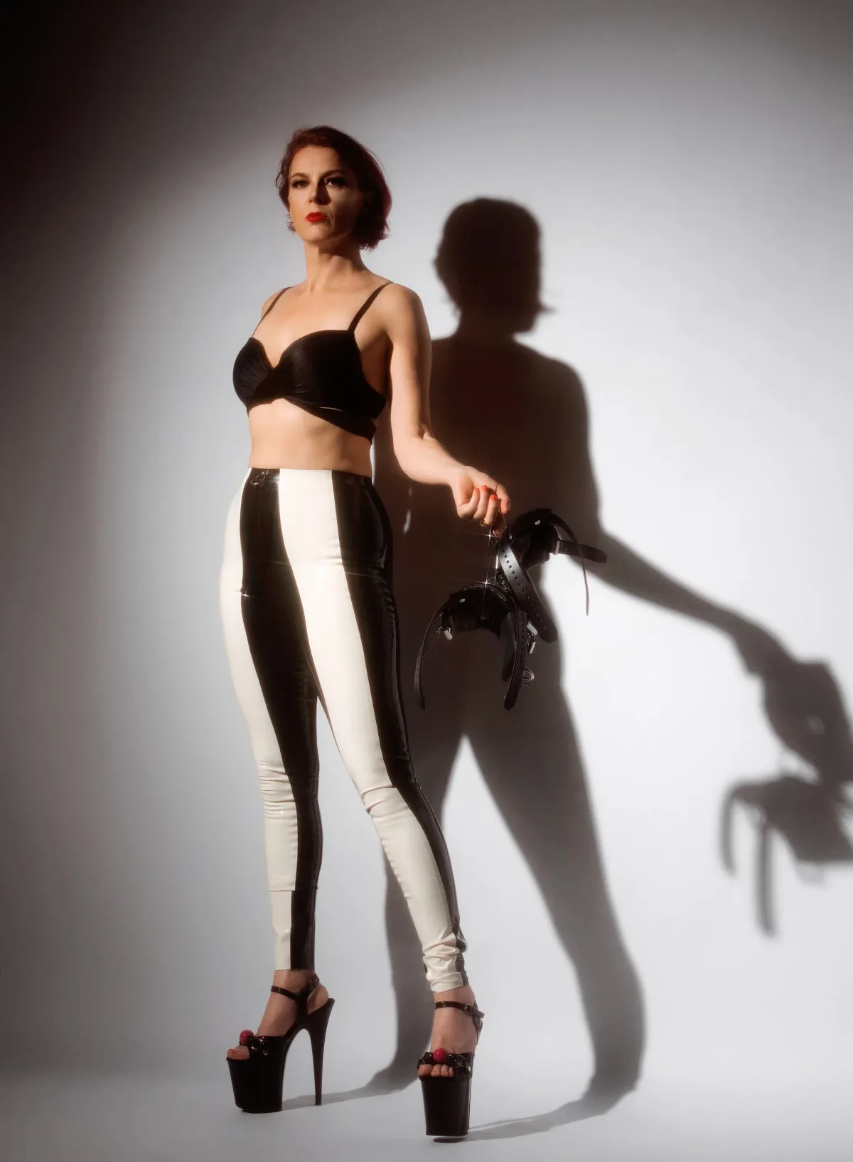 Amazon Dominatrix NYC — Goddess Viktoria Sway in latex leggings and platform heels, holding a leather harness against a dramatic shadow.