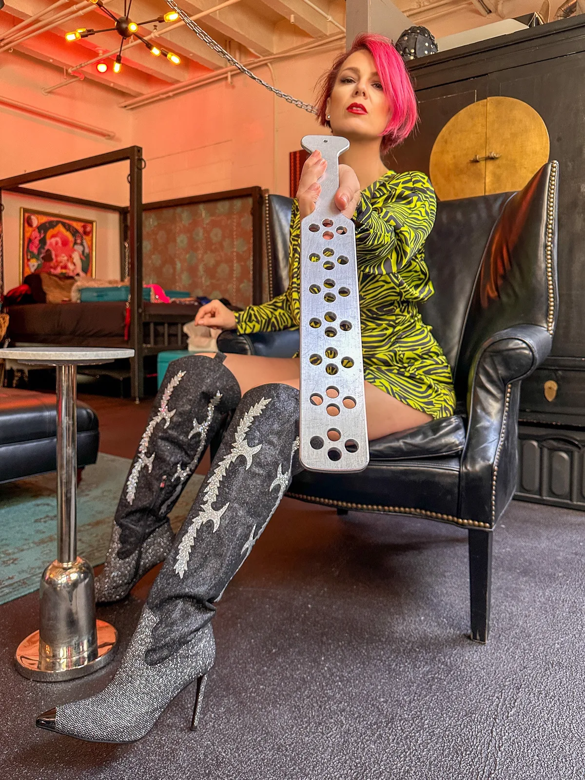 NYC Dominatrix Viktoria Sway seated in a leather wingback chair holding a metal paddle, dressed for spanking and impact play