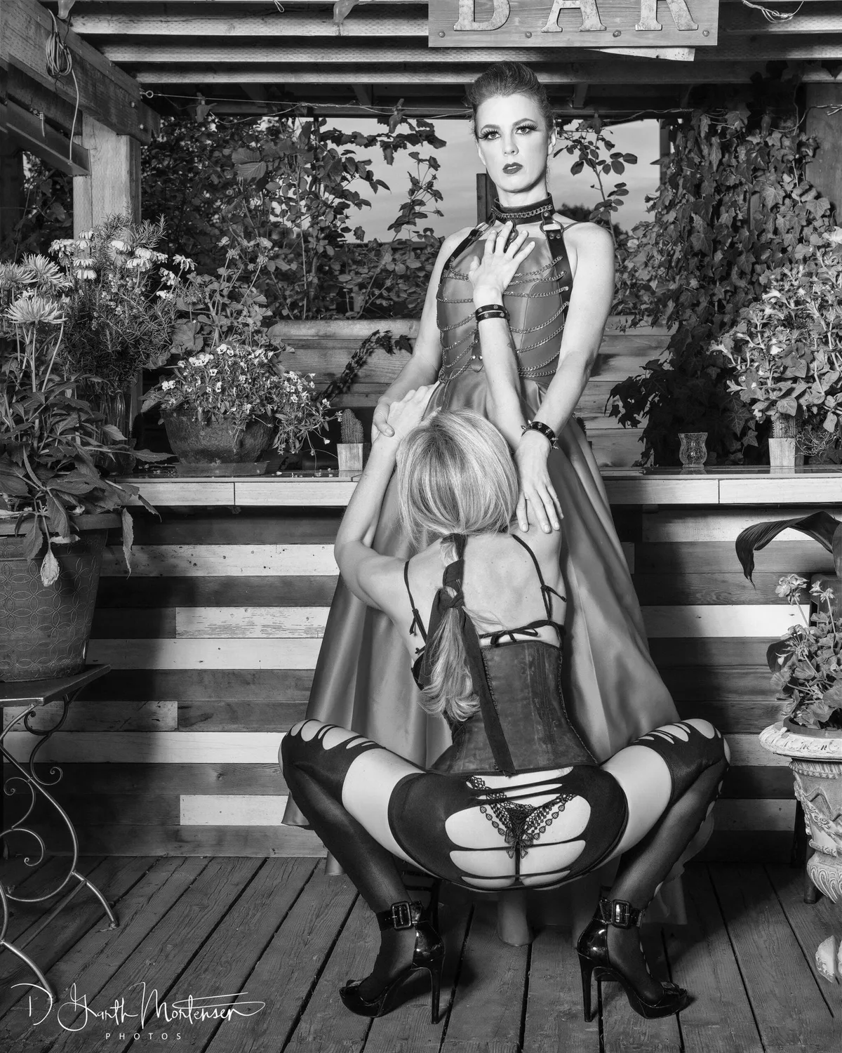 NYC Dominatrix Viktoria Sway in a chain harness and satin gown standing at a garden bar, a corseted submissive kneeling and leaning into her
